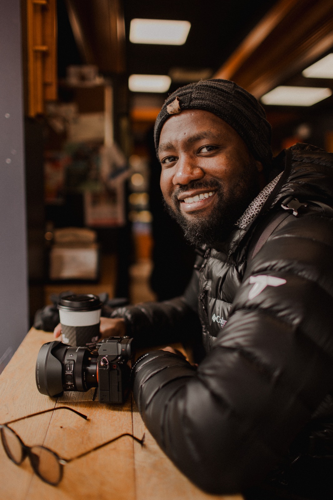 Lionel Kanyowa holding a coffee at a cafe counter, a camera resting in front of him